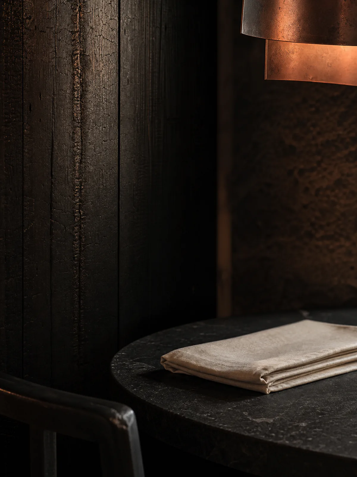 Charred timber, black stone, folded linen and a softly glowing copper light.