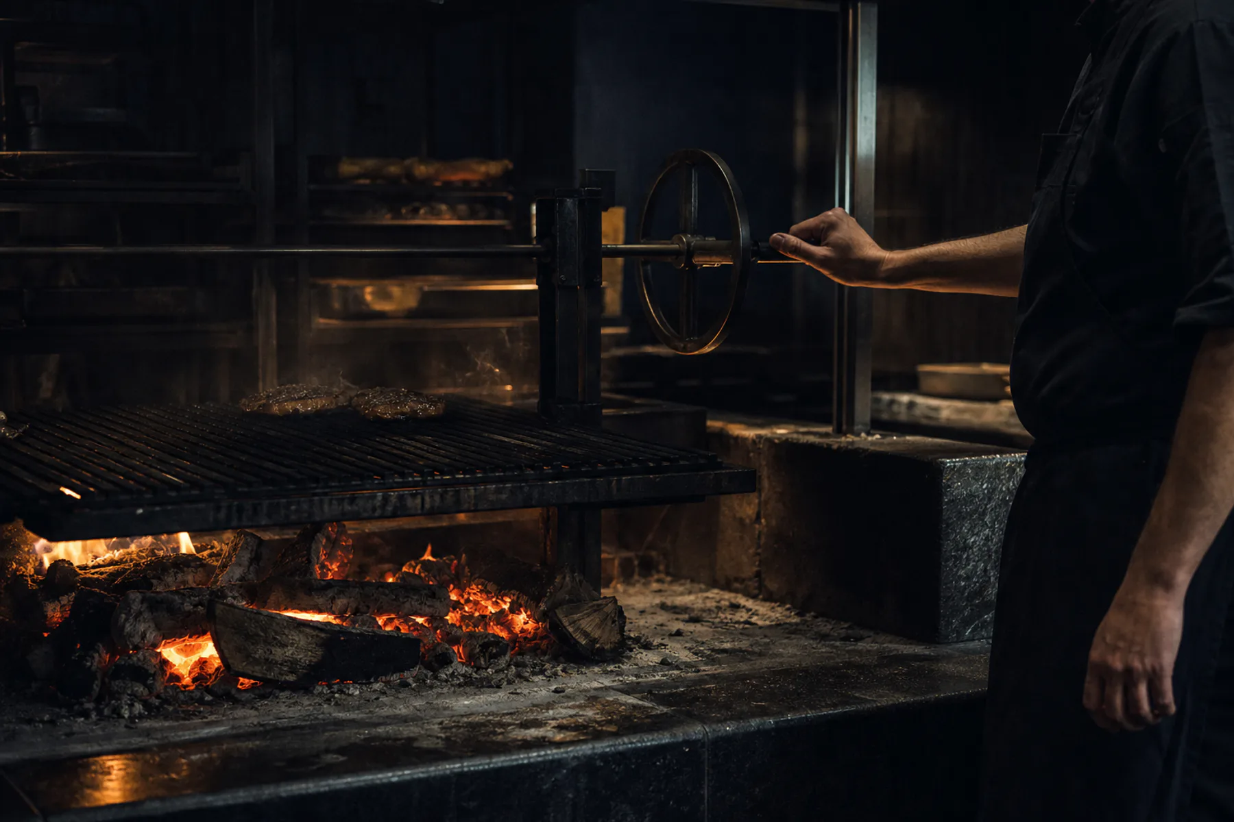 A cook adjusting the grill above glowing embers in the open kitchen.
