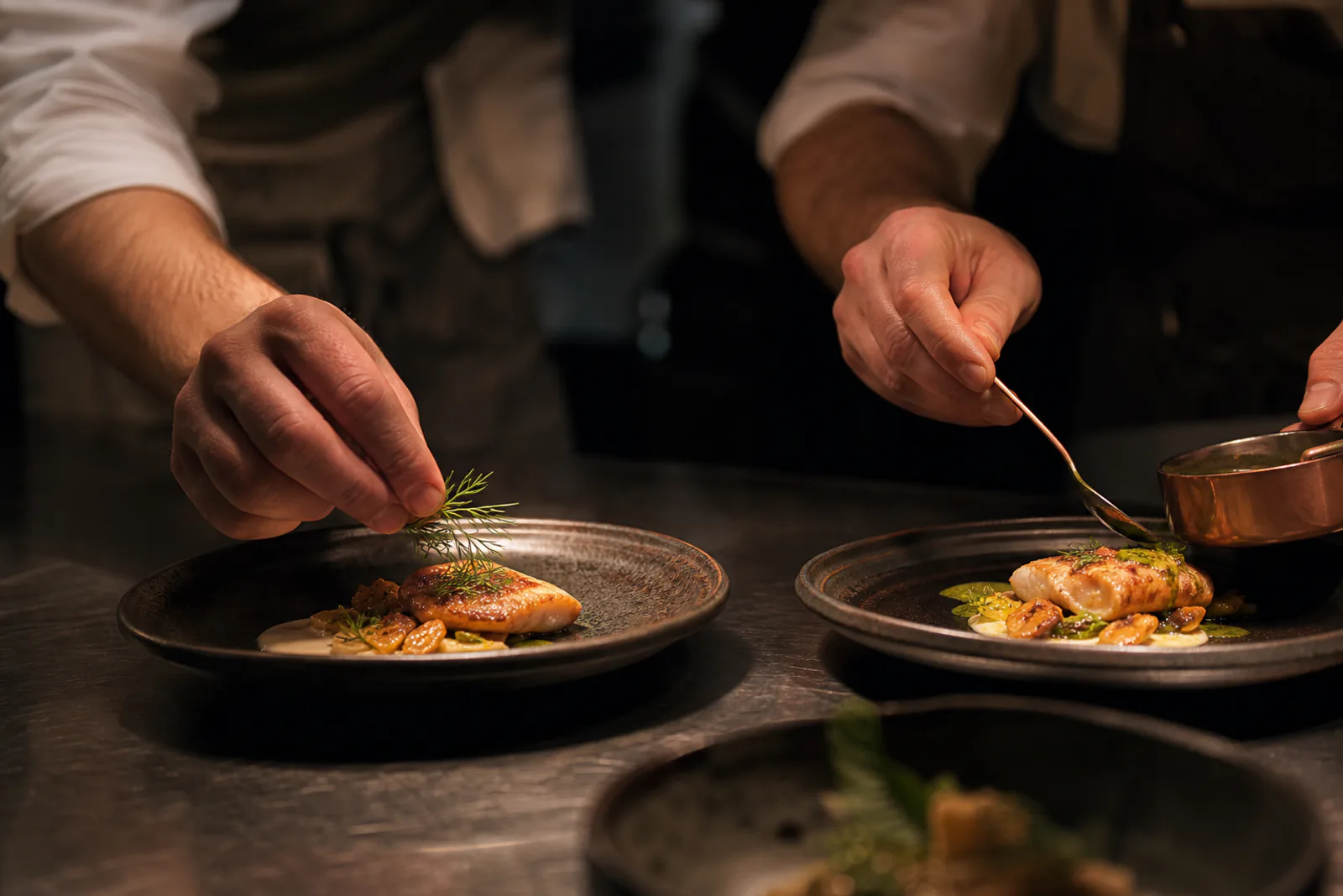 Chefs finishing dishes with herbs and sauce at the kitchen pass.